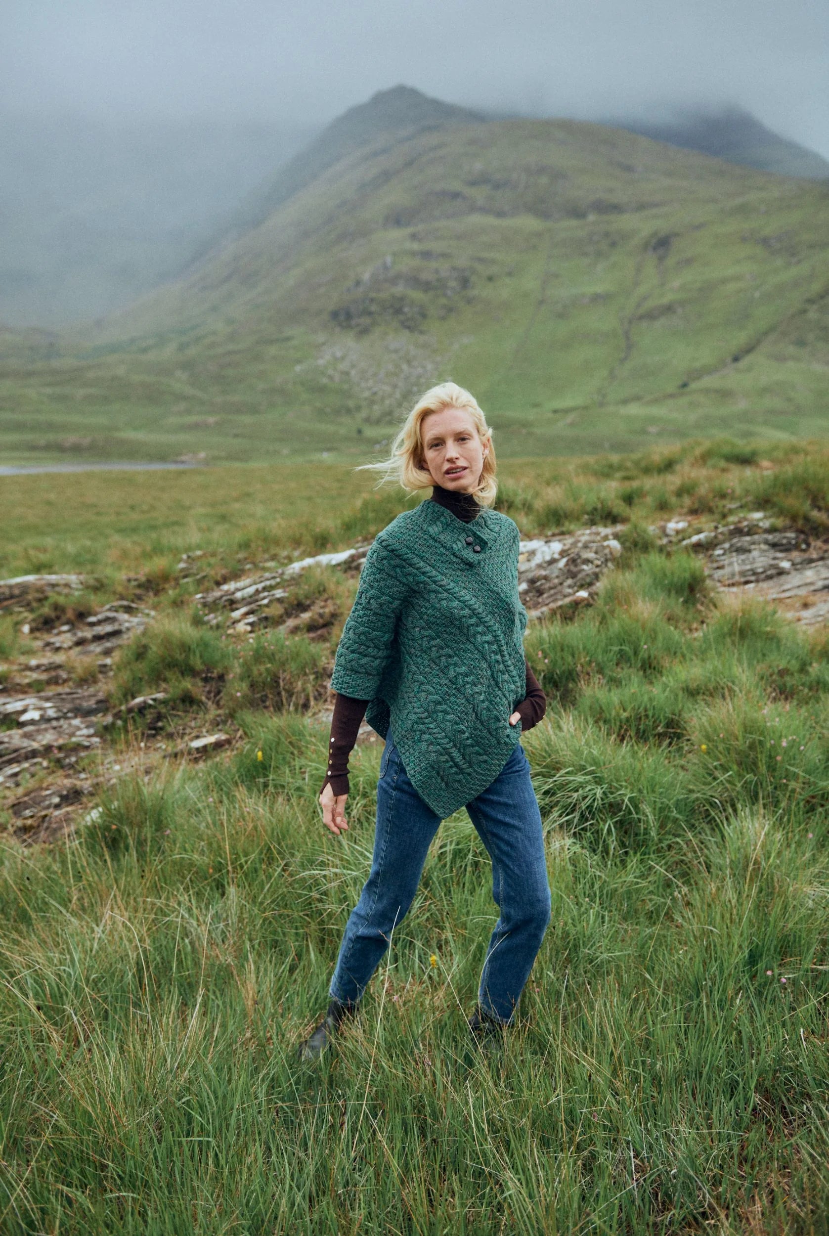 Woman wearing Easkey Ladies Merino Wool Aran button poncho in Connemara green, standing outdoors in a rugged Irish landscape.