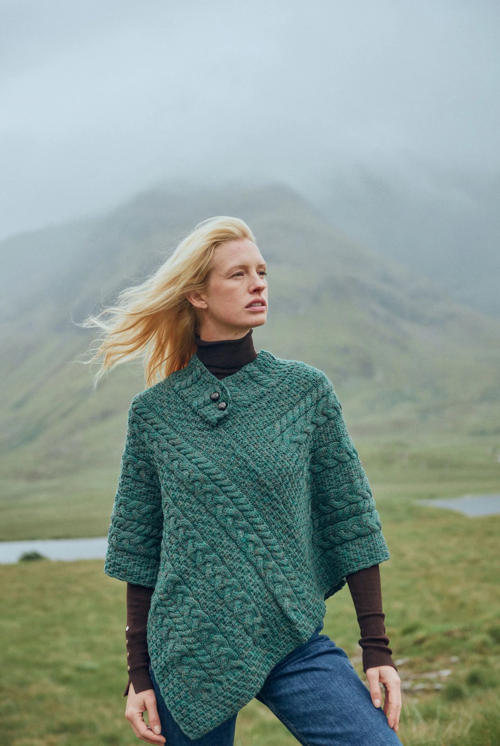 Woman wearing Easkey Connemara green Aran poncho, in the rugged Irish landscape. 

