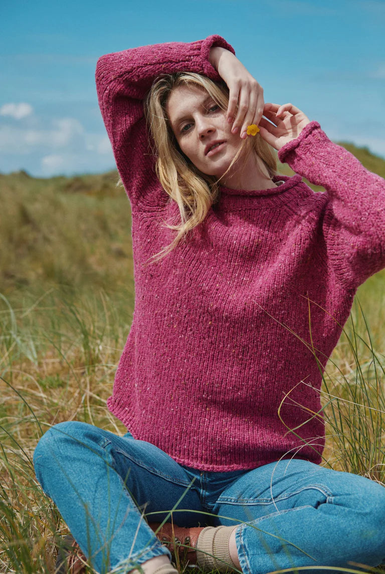 Model wearing Brackloon Tweed Roll Neck Aran Sweater in pink heather, hands raised, casual outdoor style.